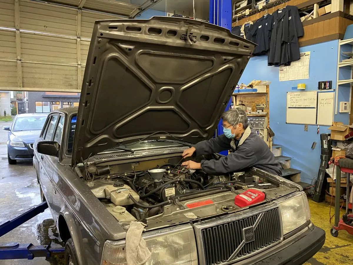 A technician working under the hood of a gray Volvo 240 in the All Vovo service bay