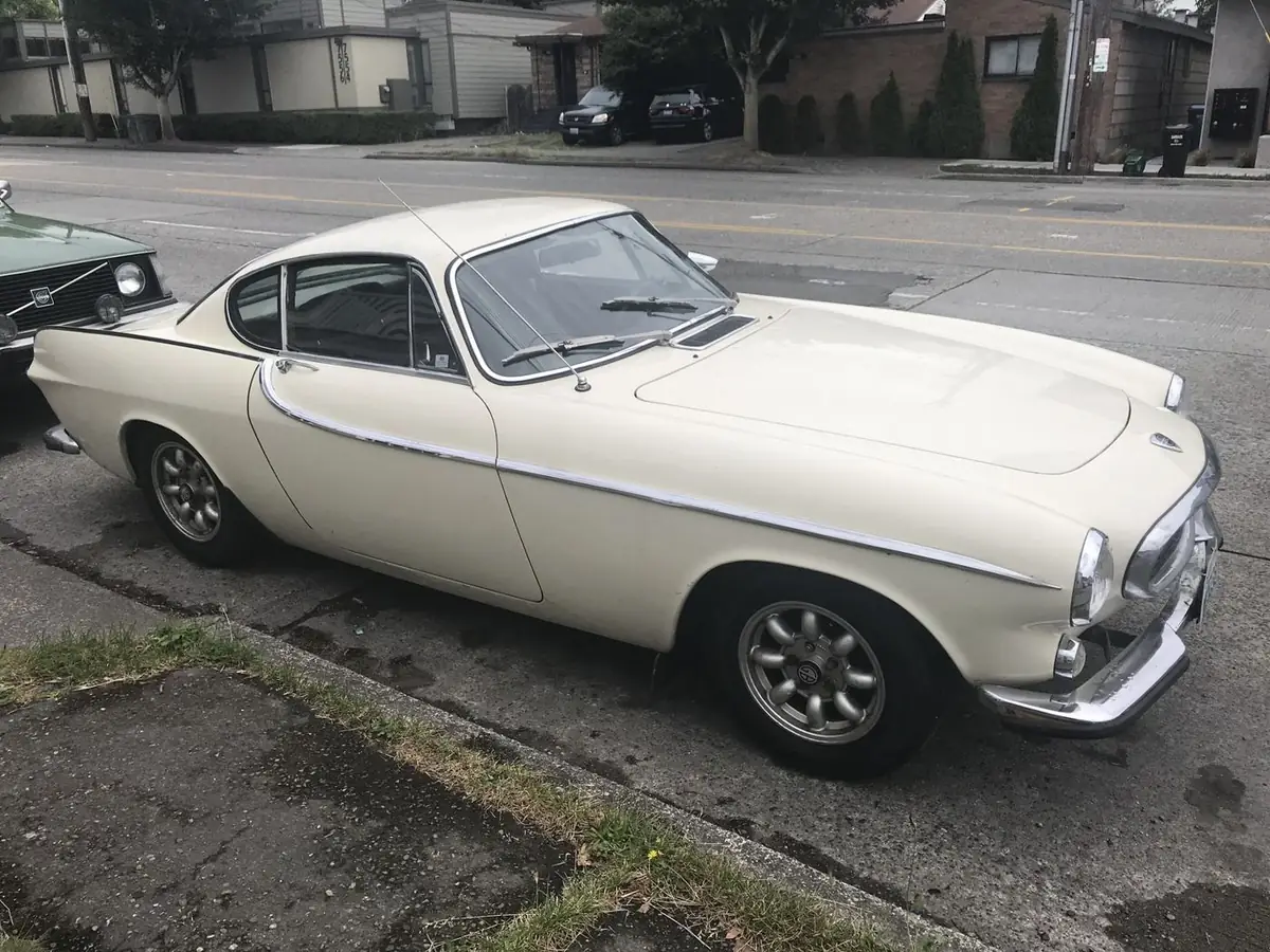 A cream Volvo P1800 coupe parked at the curb
